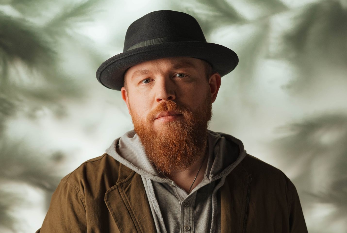 A man with a ginger beard wearing a black hat, grey jumper and green jacket